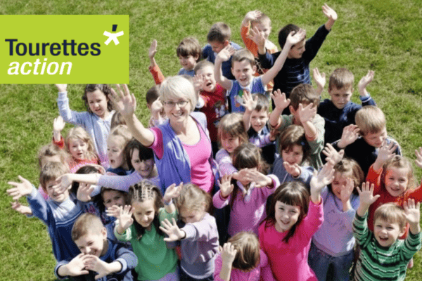 A group of children and adults outdoors, raising hands in support of Tourettes Action.