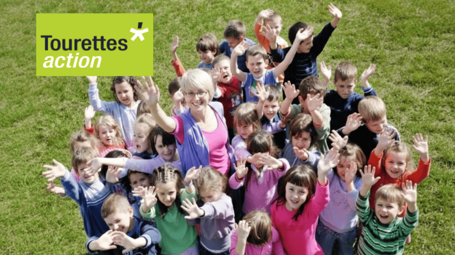 A group of children and adults outdoors, raising hands in support of Tourettes Action.
